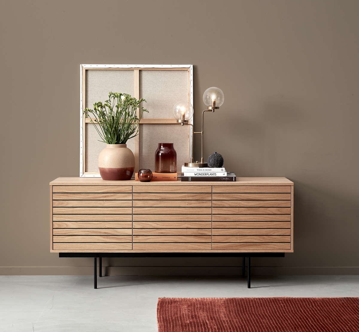 Træskænk mod beige væg med vase og grøn plante, bordlampe med glaskugle, bøgerne “This Is Home” og “Wonderplants”, glasvaser og et indrammet lærred; rustrød måtte på gulvet.