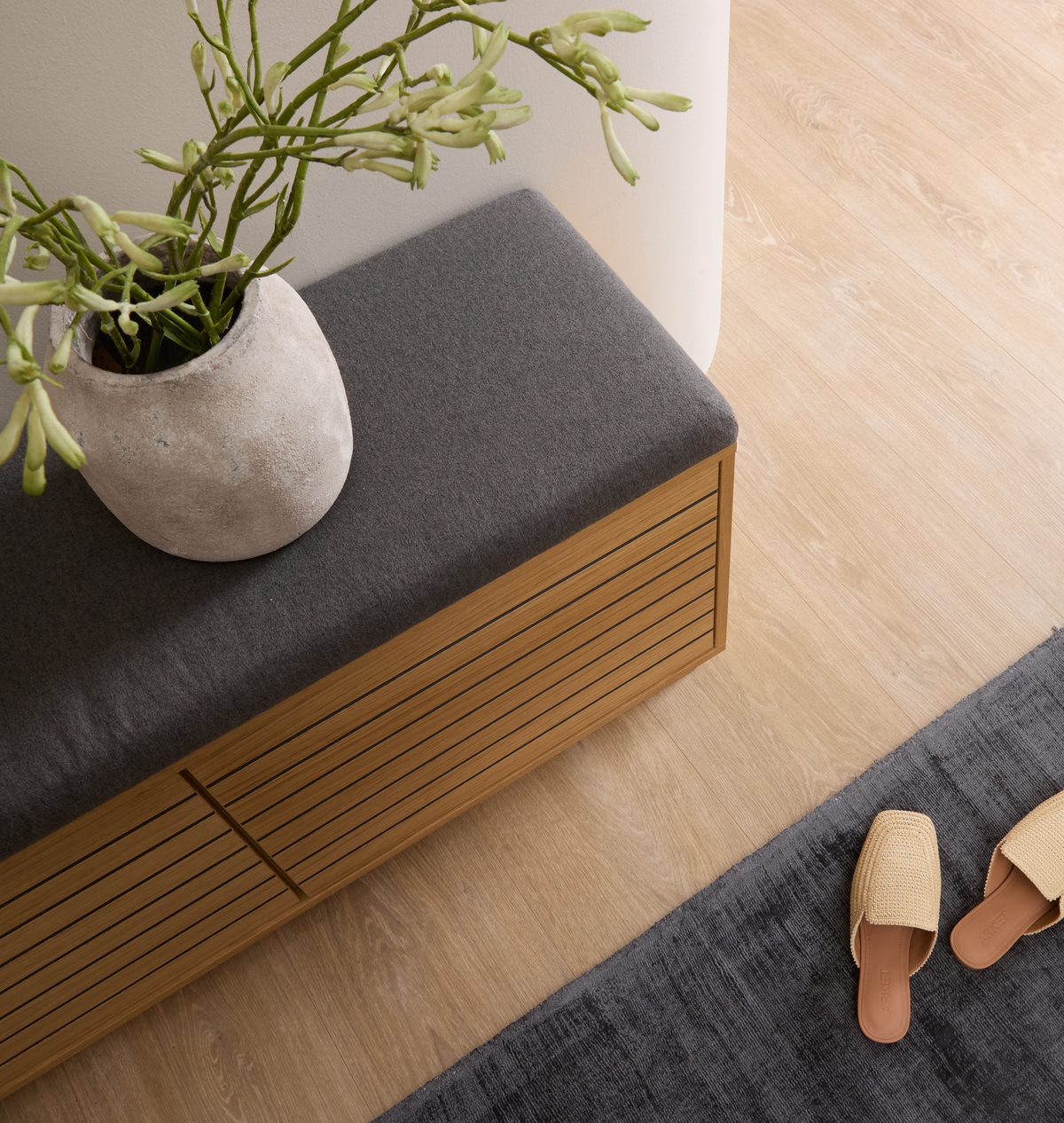 Minimalistisk entréscene med ribbet trækassebænk med grå pude, beige keramikvase med grønne kviste, mørkegråt tæppe og et par flettede slippers på lyst trægulv.