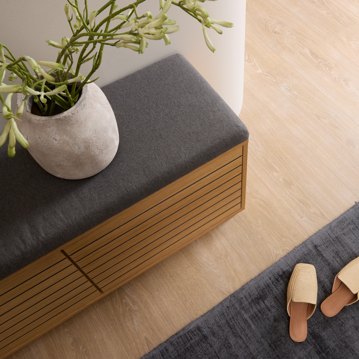 Træbænk med grå pude, keramikvase med grøn plante og et par flettede slippers på mørkegråt tæppe på lyst trægulv.