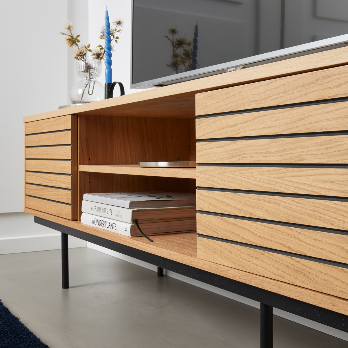 Nærbillede af et moderne mediemøbel i eg med sorte metalben; TV ovenpå, en hylde med bøgerne ”A Frame for Life” og ”Wonderplants”, samt vase med tørrede grene og et blåt snoet lys.