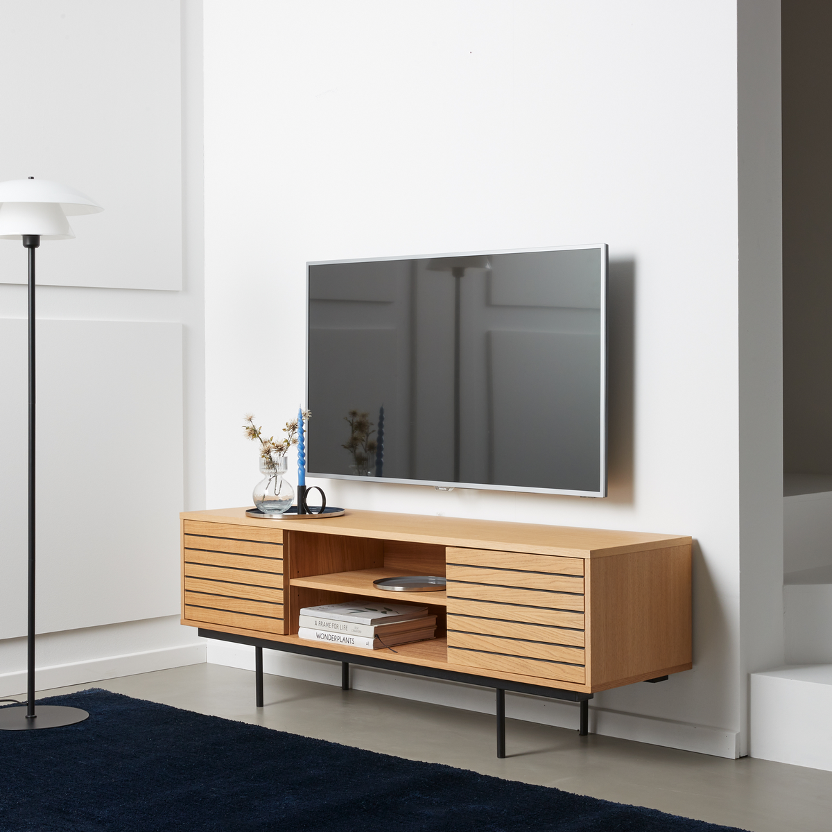 Stue med vægmonteret TV over et moderne mediemøbel i eg med lamellåger, sorte ben, blå tæppe samt detaljer som gulvlampe, vase med tørrede grene, blå snoet lys og bøgerne A Frame for Life og Wonderplants på hylden.