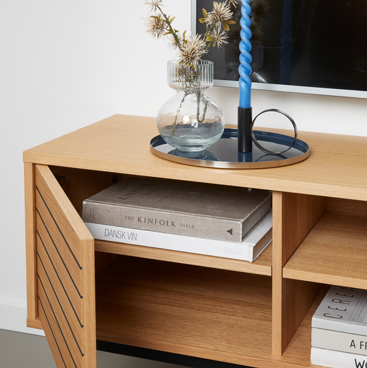 Mediemøbel i eg med glasvase med tørrede kviste, blå snoet lysestage på bakke og bøgerne The Kinfolk Table og Dansk Vin på hylden.