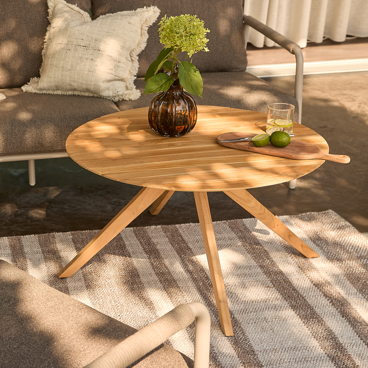 Rundt sofabord i træ på terrasse med stribet tæppe, ravfarvet vase med hortensia, skærebræt med lime og et glas; sofa i baggrunden i plettet sollys.