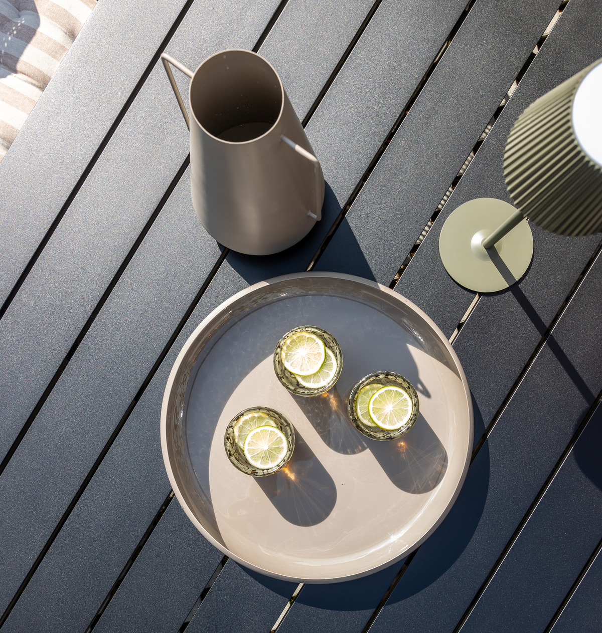 Tre glas med citronskiver på en beige bakke på et lamel-udendørsbord, med kande og lille grøn bordlampe i solskin.