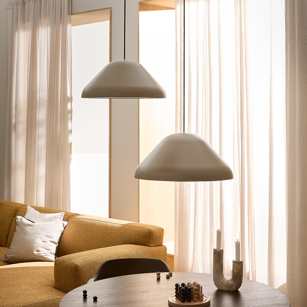 Skandinavisk stue med to beige pendellamper over rundt spisebord, sennepsgul sofa og lyse gardiner i dagslys.