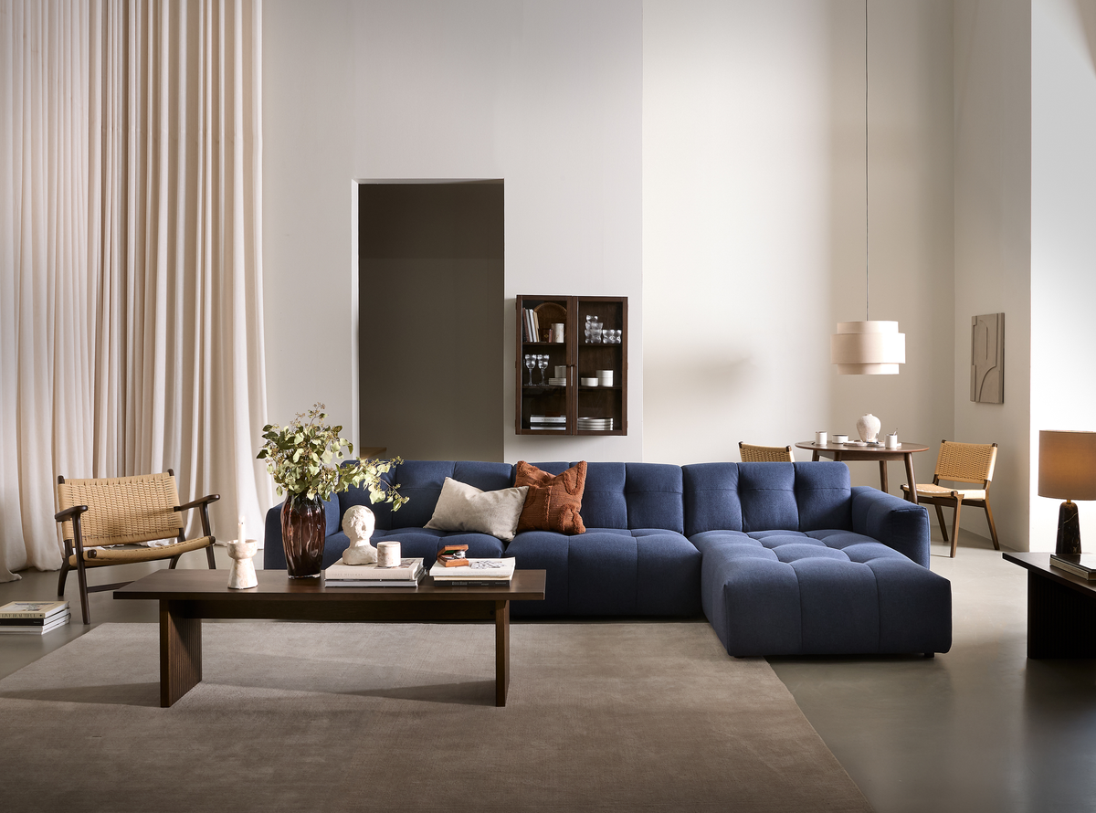 Stilrent stue-miljø med blå modulsofa, mørkt træbord med vase og bøger, beige gardiner og en pendel i en lys, neutral palet.
