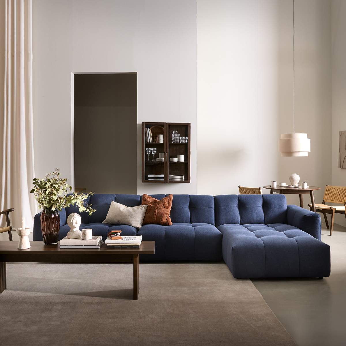 Lyst, moderne stue med mørkeblå modulsofa, træsofabord med vase og grønne grene, bøger og lys, samt lille rundt spisebord og beige pendel.