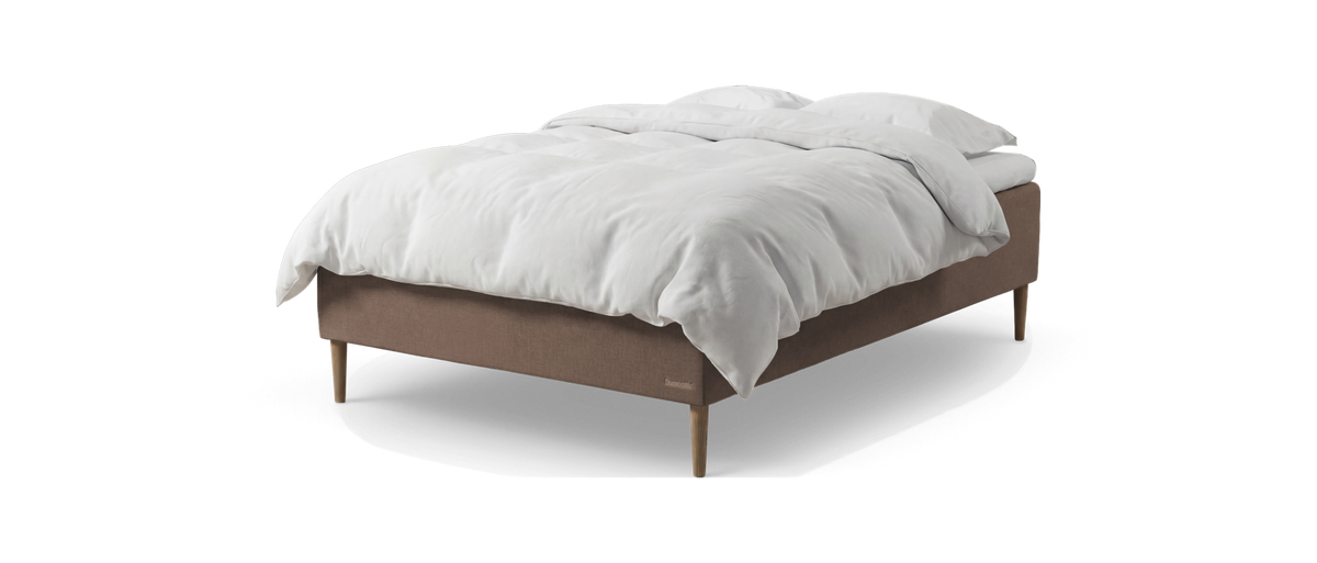 Seng med beige/brun boksmadras og træben, redt med hvid dyne og puder, frilagt på hvid baggrund.