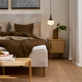 Soveværelse med Dunlopillo-seng i beige, brunt sengetøj og puder, eget natbord med vase og grøn plante, rund pendel og let gardin; bogen Mesterværker ligger på en fletbænk.