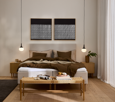 Lyst, skandinavisk soveværelse med beige seng og brunt sengetøj, to hængende pendler, fletbænk med bøger og bakke; blandt titlerne Billedkunstens historie.