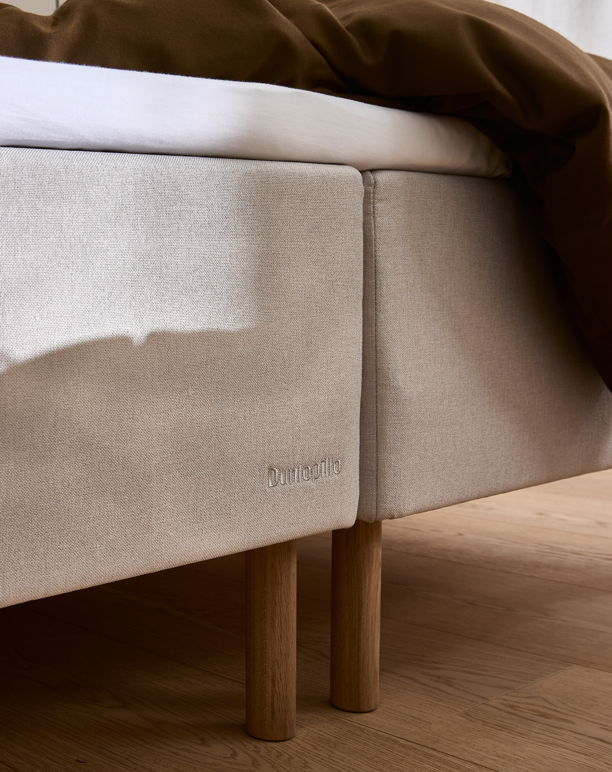 Beige tekstil-sengeramme med egetræben og hvid madras, brun dyne; Dunlopillo-branding på siden.