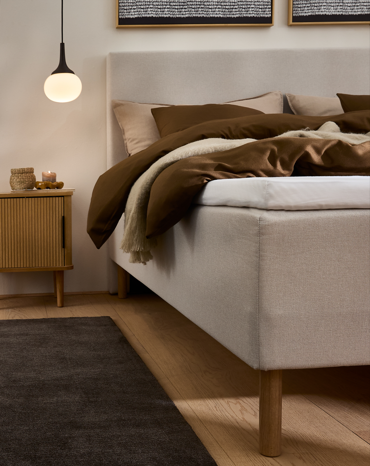 Lyst soveværelse med beige seng på træben, brunt sengetøj og plaid, rund hængende pendel, sengebord med lys og kurv samt mørkt tæppe på trægulv.