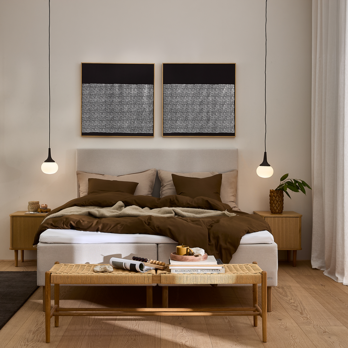 Stilrent soveværelse med beige seng, brunt sengetøj og puder, to hængende pendellamper, rottingbænk med bøger og bakke, natbord med plante og to abstrakte vægbilleder over sengegavlen.