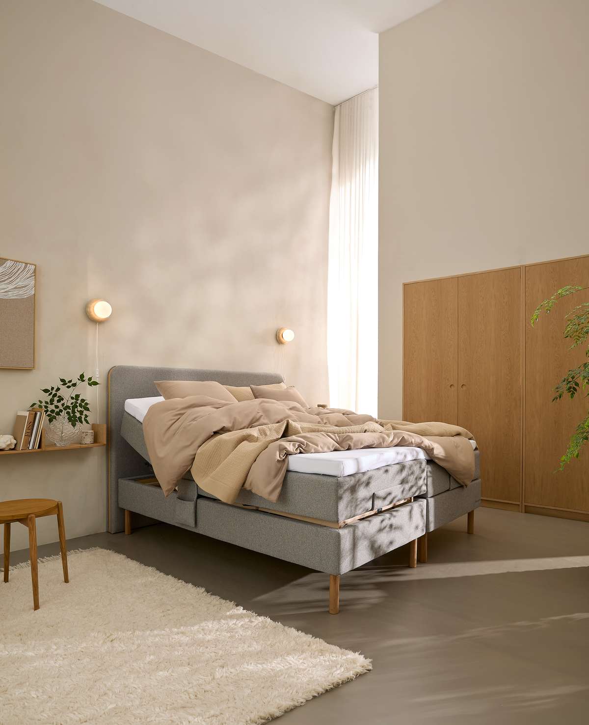 Lyst soveværelse i skandinavisk stil med en justerbar seng hvor den ene side er hævet, beige sengetøj, væglamper, træskab, hvidt tæppe og dagslys gennem lette gardiner.