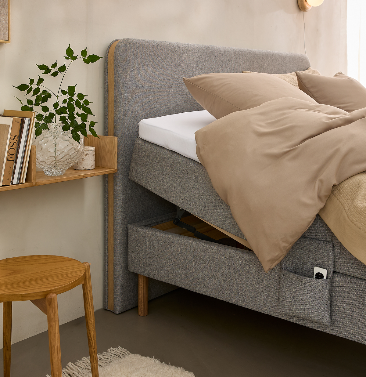 Grå polstret seng med løftet madras, beige sengetøj og puder; egetræsskub, hylde med glasvase, grøn plante og bøger inklusive titlen BOSS, blødt tæppe og væglampe i et lyst soveværelse.