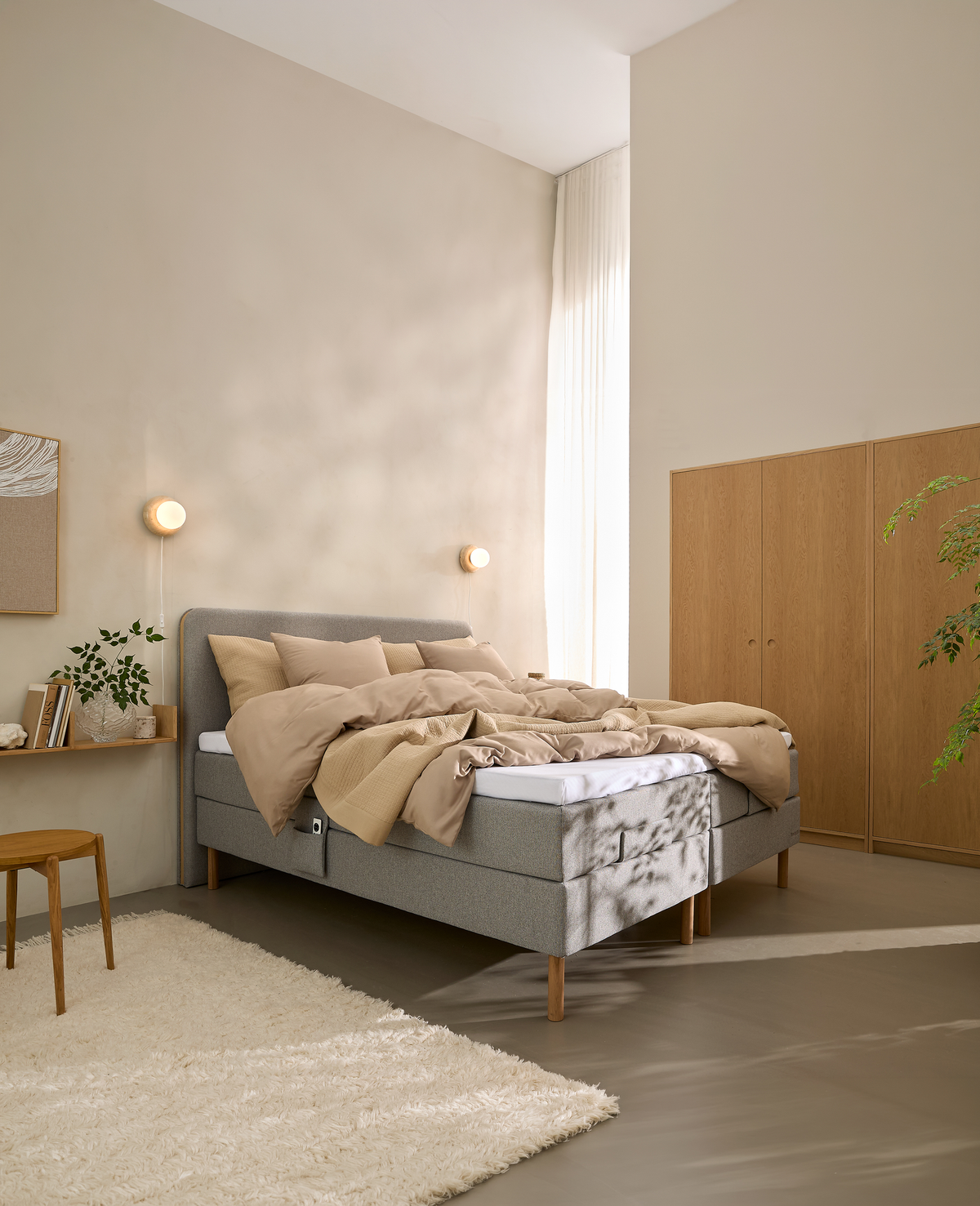 Lyst soveværelse med grå kontinentalseng, beige sengetøj, vægmonterede lamper, lyse gardiner og garderobeskab i træ.