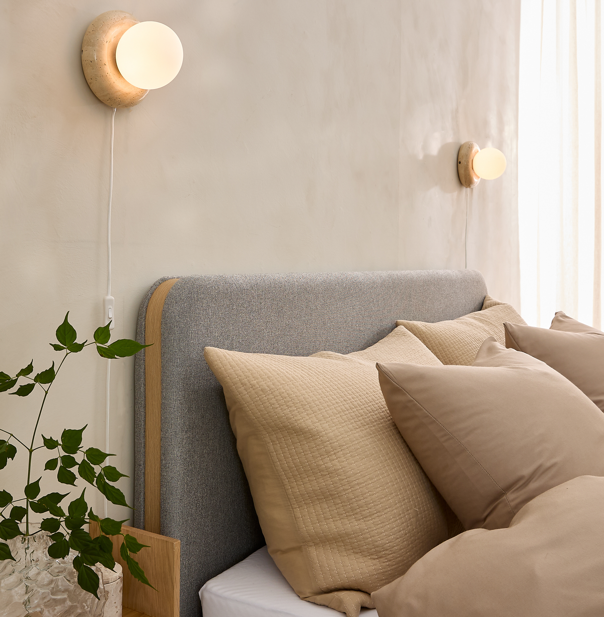 Seng med grå polstret sengegavl og beige puder, to tændte runde væglamper og dagslys gennem lyse gardiner; grøn plante på sengebord.