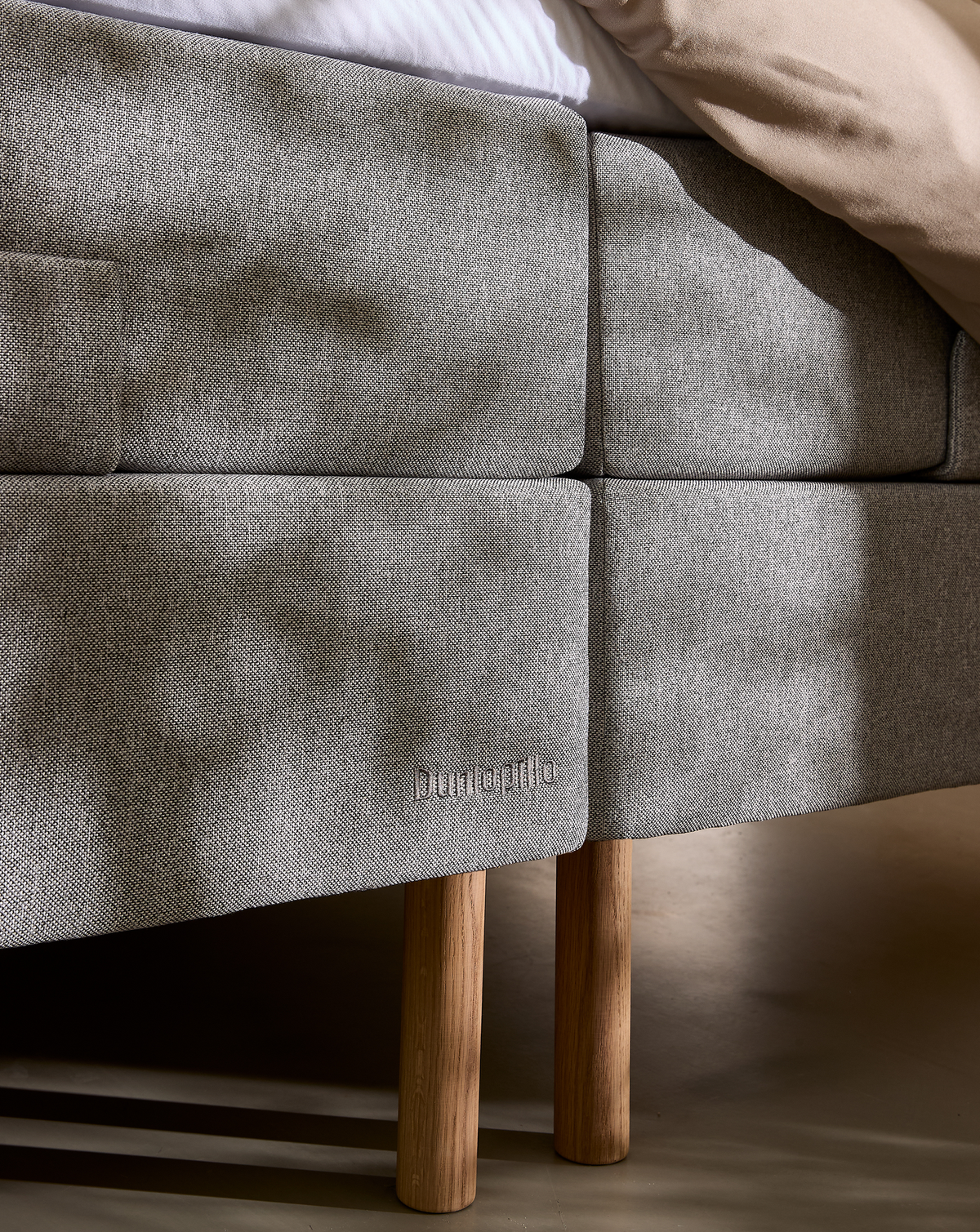 Gråpolstret sengebund med træben, Dunlopillo‑branding, bløde dagslysskygger og beige dyne.