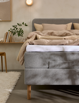 Grå polstret seng med beige sengetøj i roligt soveværelse med væglampe, hylde med bøger og grøn plante; blød tæppe og varme neutrale toner.