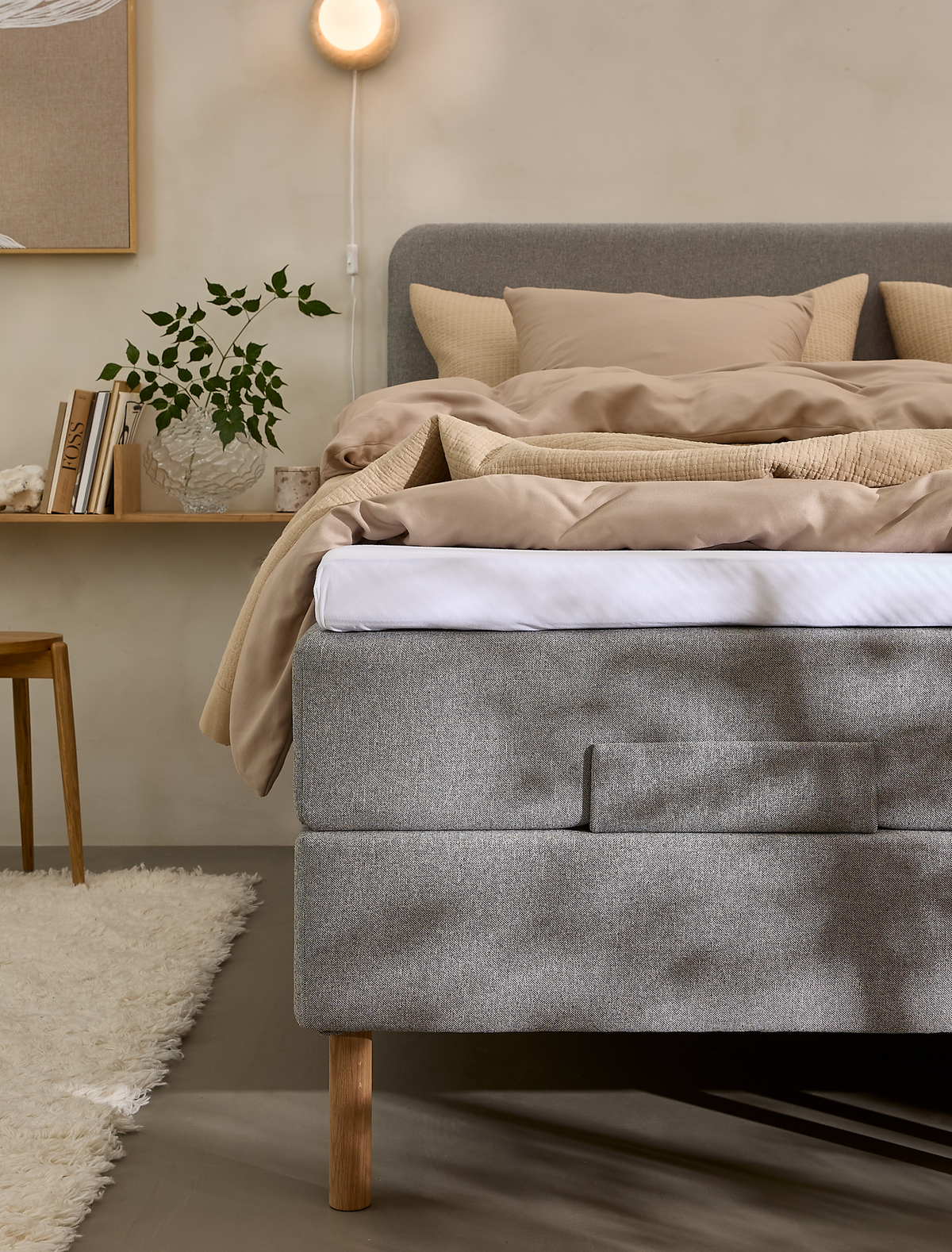 Grå polstret seng med beige sengetøj i roligt soveværelse med væglampe, hylde med bøger og grøn plante; blød tæppe og varme neutrale toner.