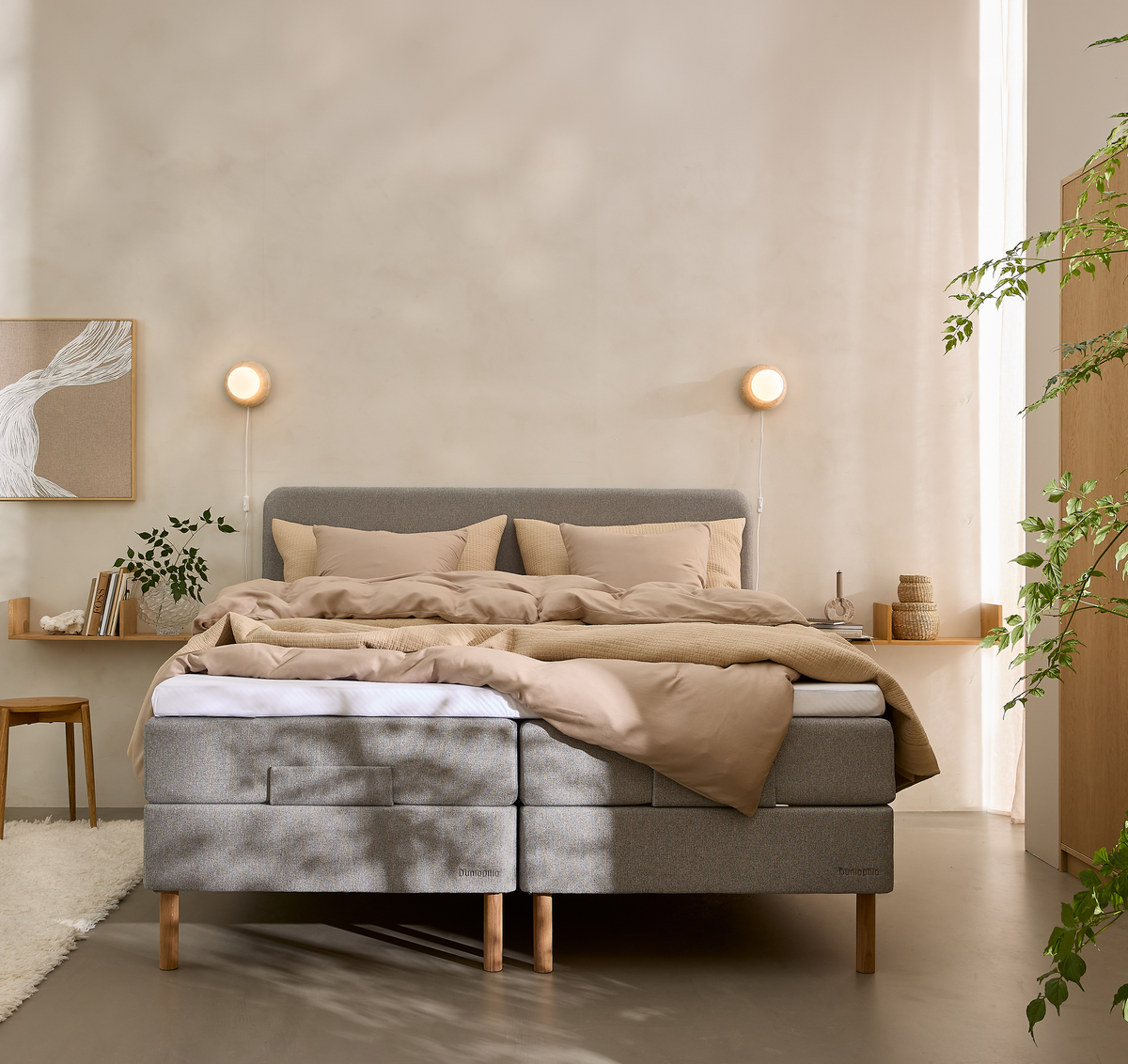 Lyst soveværelse med grå kontinentalseng, beige sengetøj, væglamper og planter.