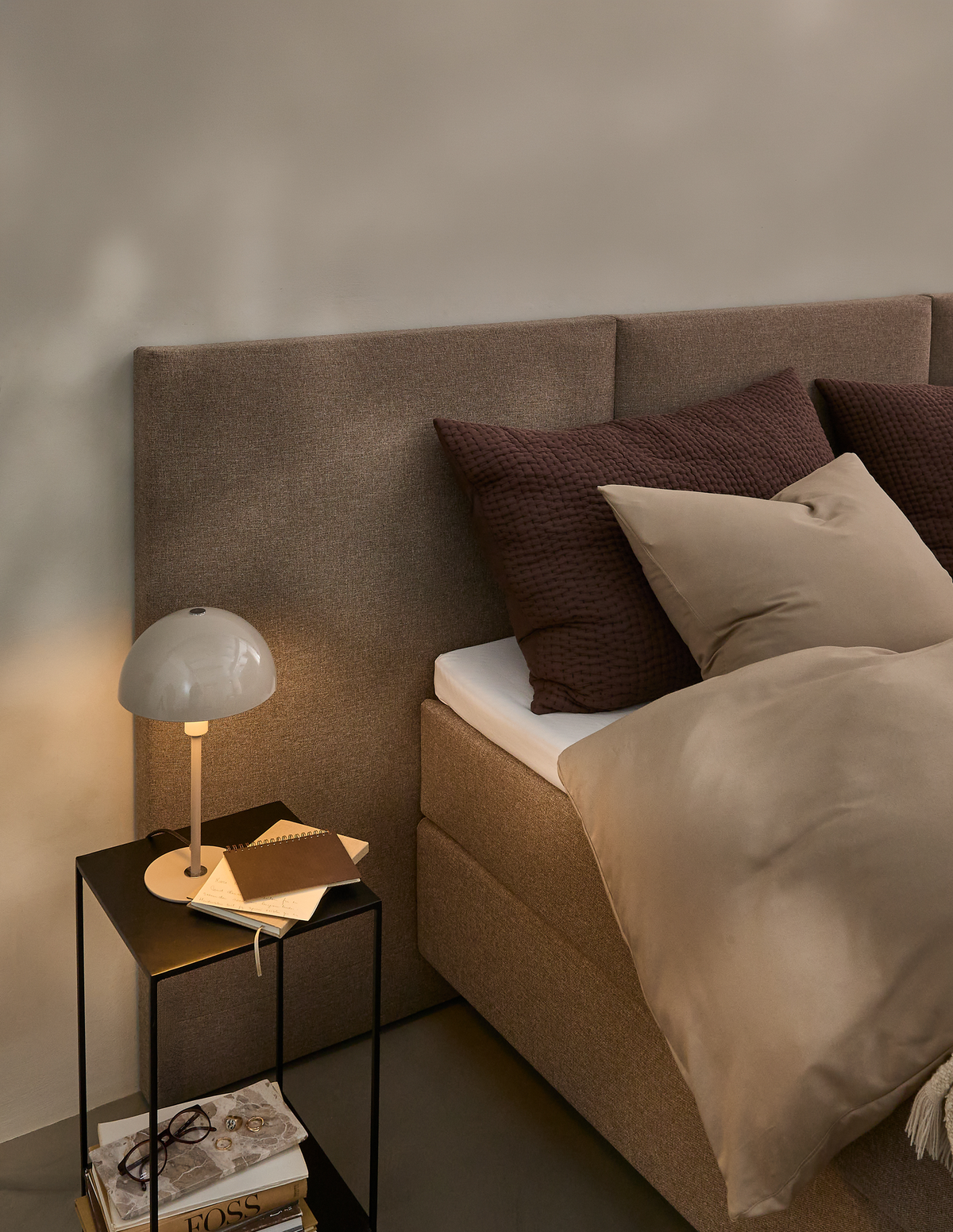 Soveværelseshjørne med beige seng, brune puder og et lille sort sidebord med bordlampe og bøger.