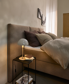 Soveværelse med polstret sengegavl, beige sengetøj og sengebord med bordlampe, bøger og briller.