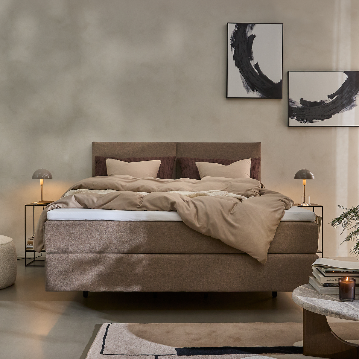 Beige polstret seng med neutralt sengetøj i et minimalistisk skandinavisk soveværelse, sideborde med små kuppellamper, abstrakt sort-hvid vægkunst, tæppe og sofabord med lys.