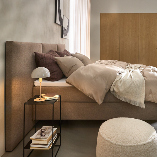 Lyst soveværelse med beige seng, lille sengebord med bordlampe og bøger og et garderobeskab i træ i baggrunden.