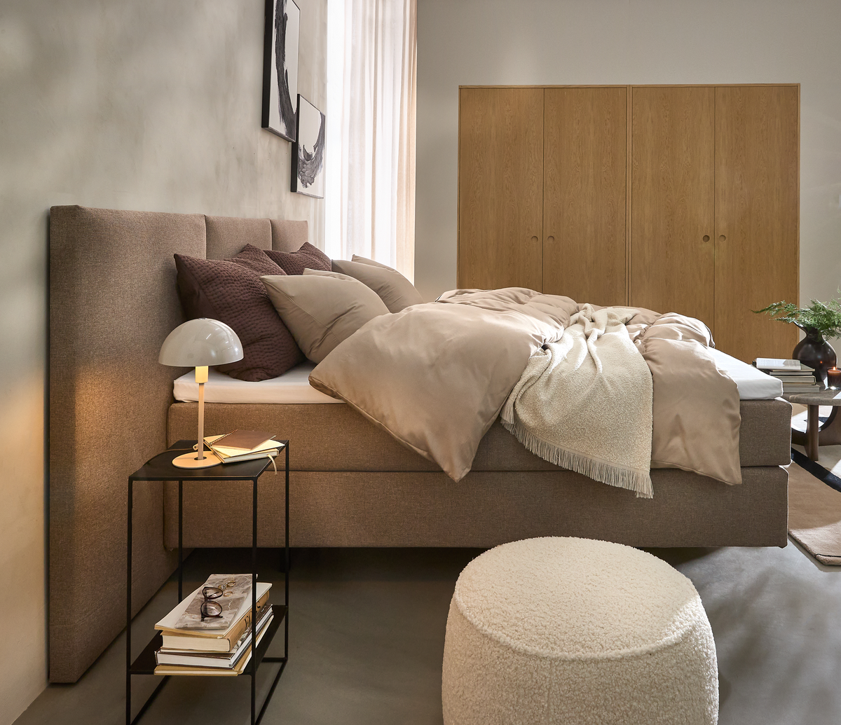Skandinavisk soveværelse med beige polstret seng, lag af puder og dyne, sengebord med lille lampe og bøger, lys træskabsvæg, rund puf og grøn plante i blødt dagslys.