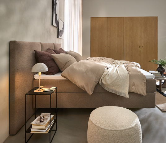 Skandinavisk soveværelse med beige polstret seng, lag af puder og dyne, sengebord med lille lampe og bøger, lys træskabsvæg, rund puf og grøn plante i blødt dagslys.