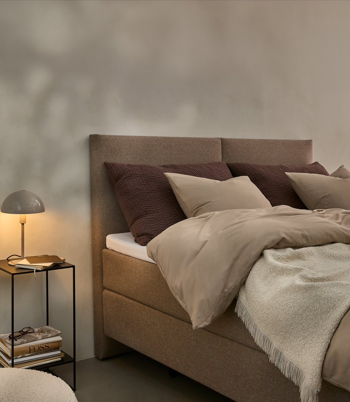 Soveværelse med beige seng med brune og beige puder, beige dyne og lys plaid; sort sengebord med bordlampe og stablede bøger ved siden af.