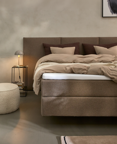 Beige og brun soveværelsesscene med polstret kontinentalseng, puder og dyner, natbord med bordlampe, rund puf og lille tæppe.