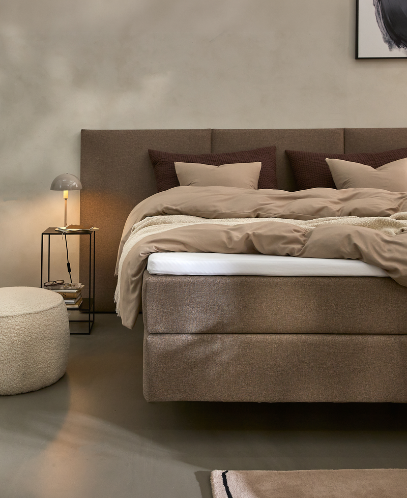 Beige og brun soveværelsesscene med polstret kontinentalseng, puder og dyner, natbord med bordlampe, rund puf og lille tæppe.