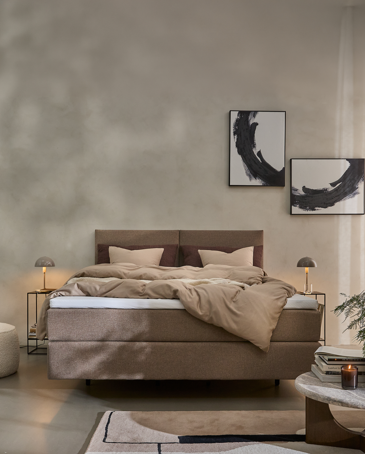 Lyst og minimalistisk soveværelse med beige kontinentalseng, symmetriske sengeborde med små lamper, abstrakt sort-hvid vægkunst og lavt bord med bøger og tændt lys.