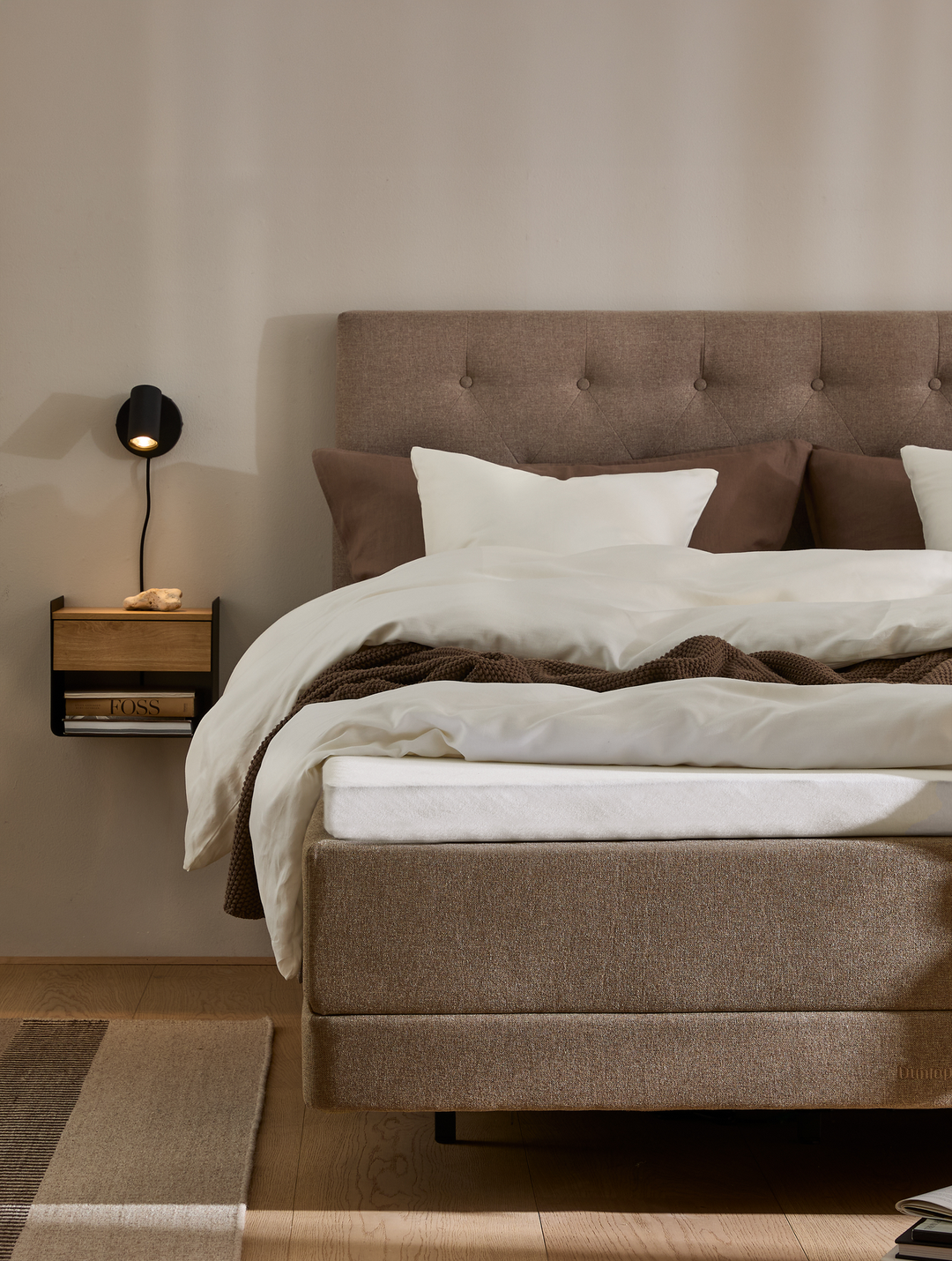 Neutralt soveværelse med polstret sengegavl i beige, hvide og brune sengelinned, vægmonteret sort læselampe og lille hylde med bog; varm, minimalistisk stil.