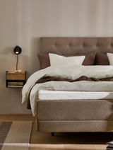 Neutralt soveværelse med polstret sengegavl i beige, hvide og brune sengelinned, vægmonteret sort læselampe og lille hylde med bog; varm, minimalistisk stil.