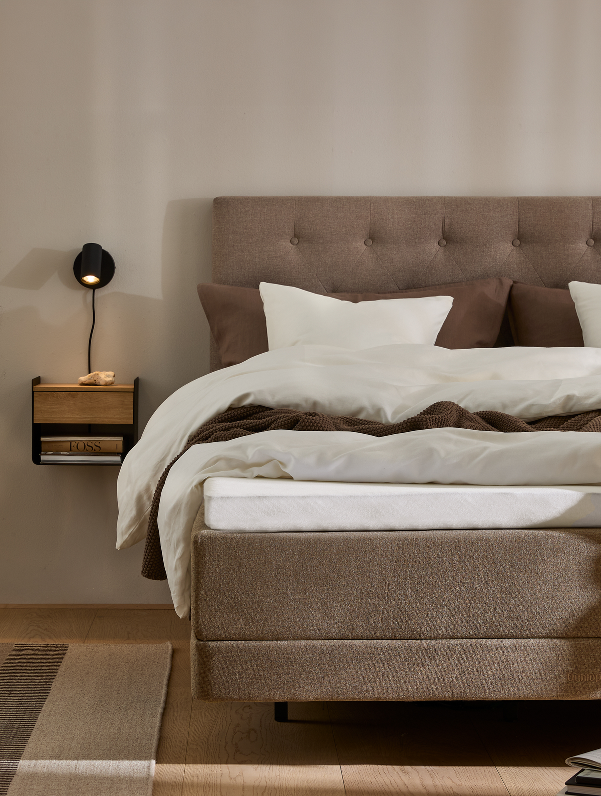 Neutralt soveværelse med polstret sengegavl i beige, hvide og brune sengelinned, vægmonteret sort læselampe og lille hylde med bog; varm, minimalistisk stil.
