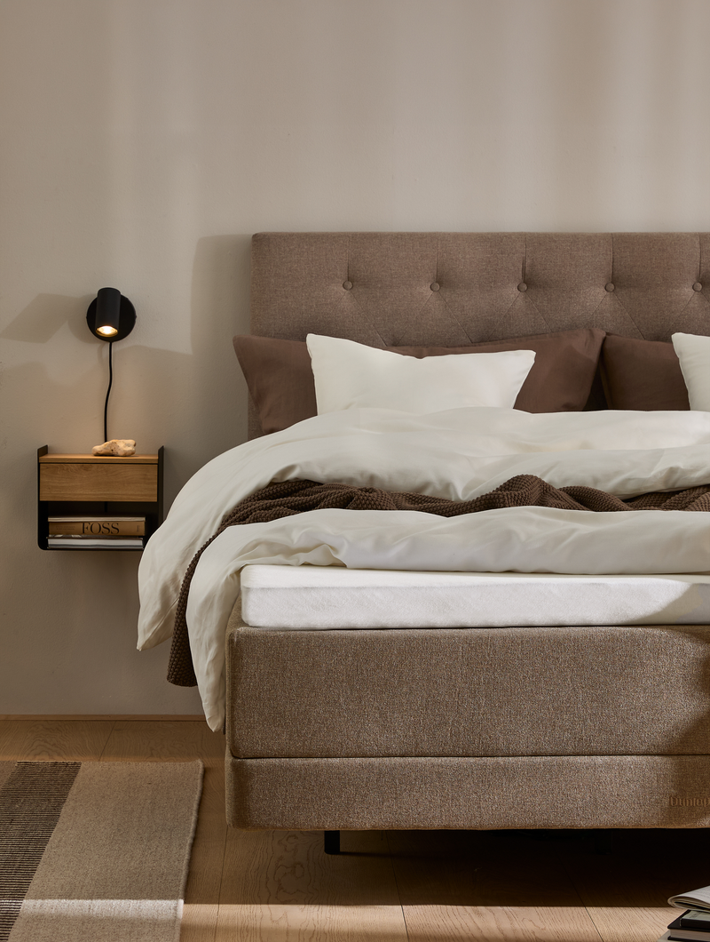 Neutralt soveværelse med polstret sengegavl i beige, hvide og brune sengelinned, vægmonteret sort læselampe og lille hylde med bog; varm, minimalistisk stil.