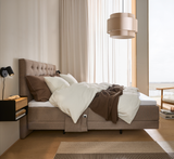 Lyst soveværelse med polstret kontinentalseng i beige, hvide og brune tekstiler, brun plaid, beige pendel, sort væglampe, lyse gardiner og et tæppe i naturtoner ved et stort vindue.
