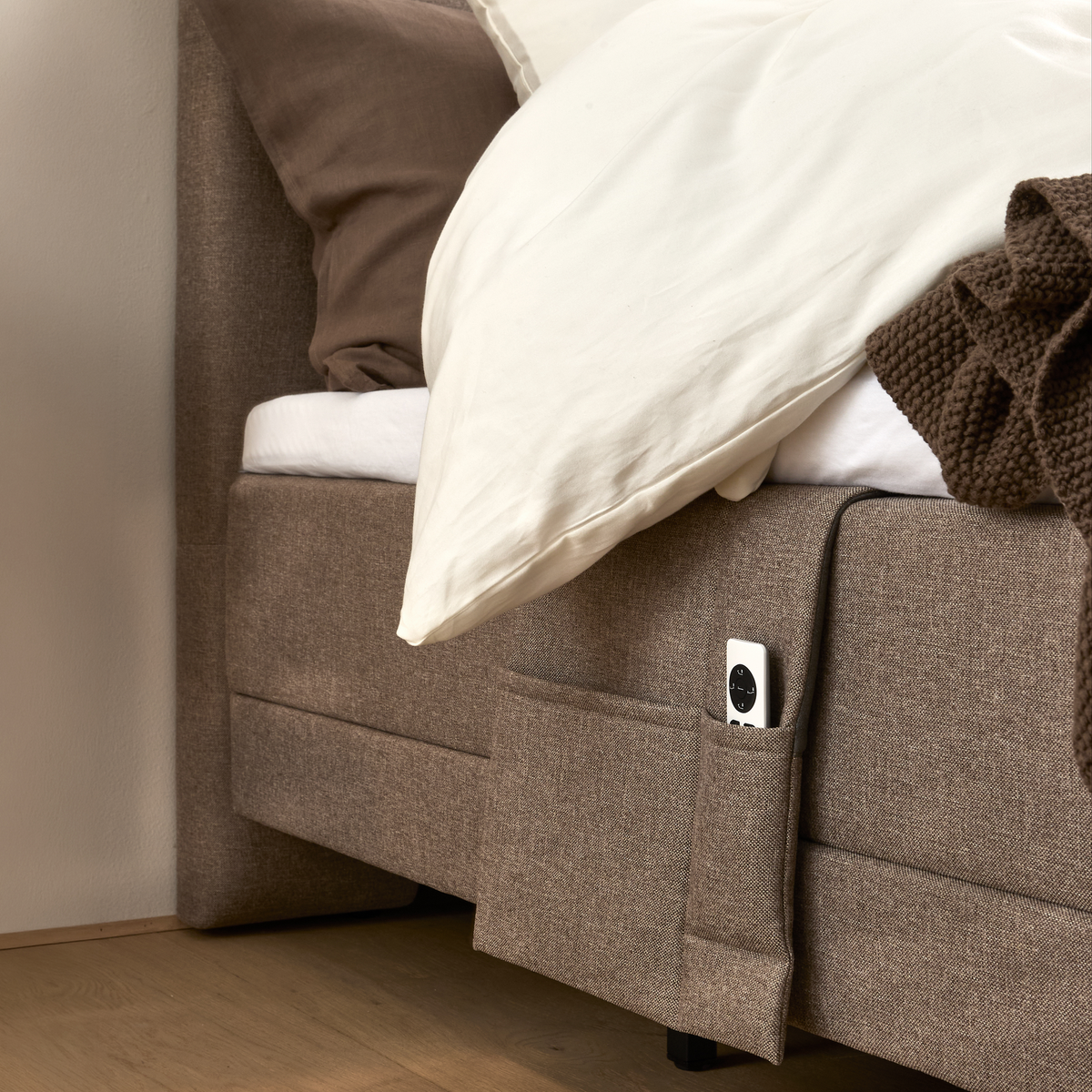 Seng med stoflomme på siden med en fjernbetjening, puder og strikket plaid i beige nuancer.