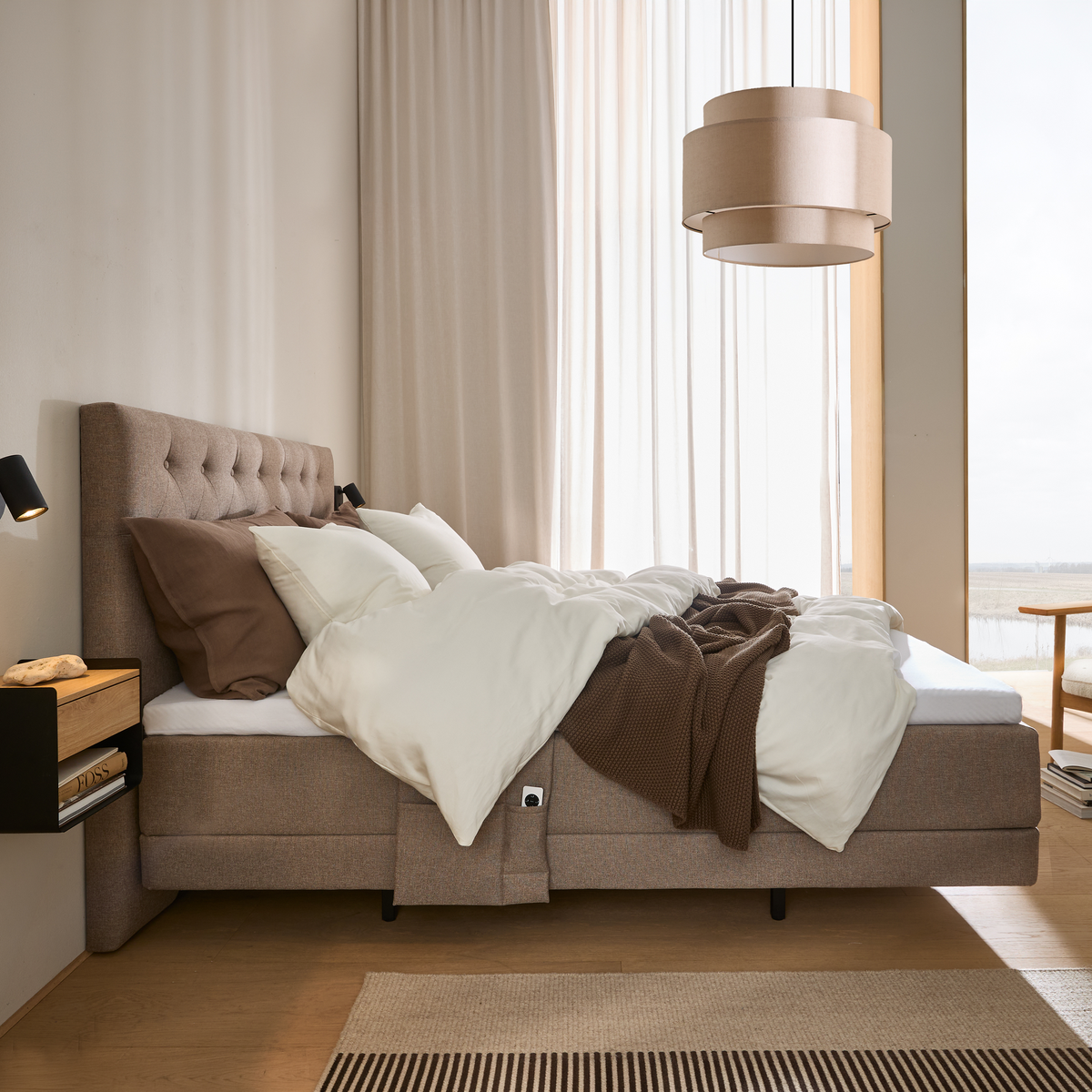Beige soveværelse med polstret seng, hvide dyner og brun plaid ved stort vindue med lyse gardiner; beige pendel og vægmonteret sengebord.