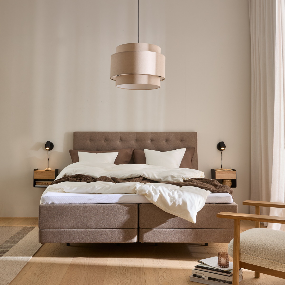 Lyst, neutralt soveværelse i skandinavisk stil med polstret seng, beige pendel, svævende sengeborde med små sorte væglamper og hvid/brun sengetøj.