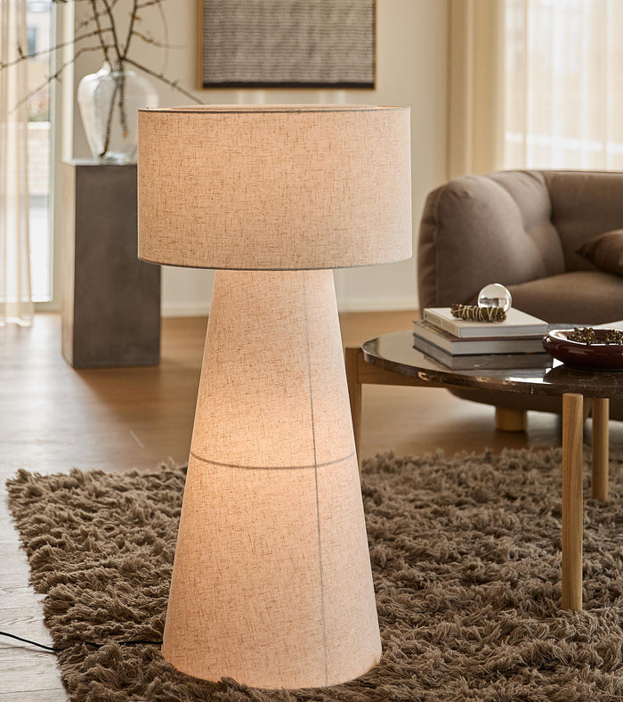 Beige gulvlampe med konisk, stofbeklædt fod og stor cylindrisk skærm i en skandinavisk stue med ryatæppe og sofabord.
