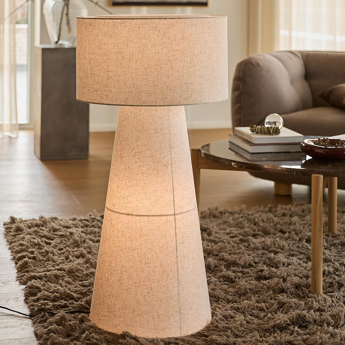 Beige gulvlampe med konisk, stofbeklædt fod og stor cylindrisk skærm i en skandinavisk stue med ryatæppe og sofabord.