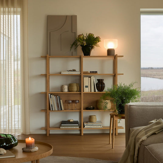 Lyst og varmt stuefoto med en reol i eg stylet med bøger, kurve, planter og en glødende bordlampe; tændt stearinlys på rundt sofabord og neutral sofa ved stort vindue.