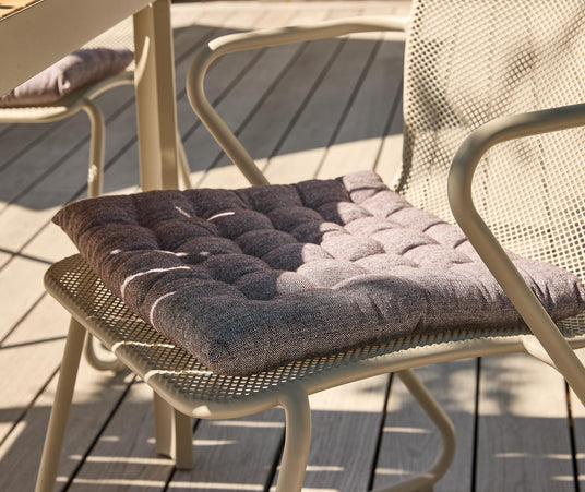 Beige metalstol på træterrasse med tuftet grå sædehynde i sollys.