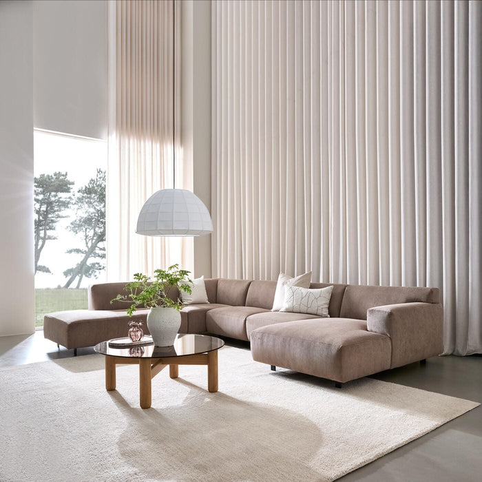 Lys stue med beige hjørnesofa, rundt sofabord i glas med vase og grøn plante, stor hvid pendel og gulvlange lyse gardiner ved et stort vindue.