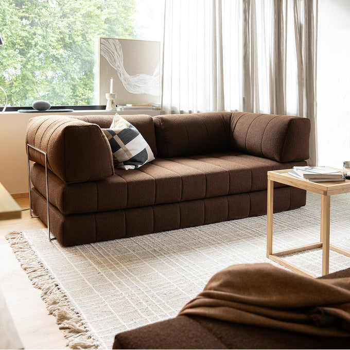 Brun modulsofa i en moderne stue med lyst, tekstureret tæppe, lille træbord og lette gardiner ved et stort vindue; sort‑hvid ternet pude på sofaen.