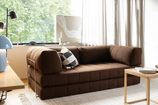 Brun modulsofa i en moderne stue med lyst, tekstureret tæppe, lille træbord og lette gardiner ved et stort vindue; sort‑hvid ternet pude på sofaen.